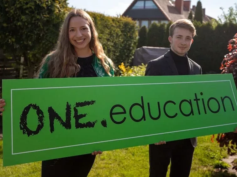 Die Gründerin Laura und der Gründer Joscha stehen im Garten und halten ein großes Schild mit dem Namen ihres Startups One.Education hoch.