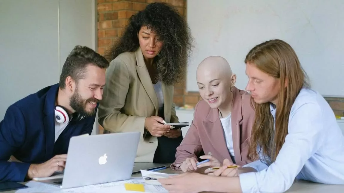 Das Bild zeigt vier diverse Personen, die gemeinsam an einem Schreibtisch sitzen und arbeiten.