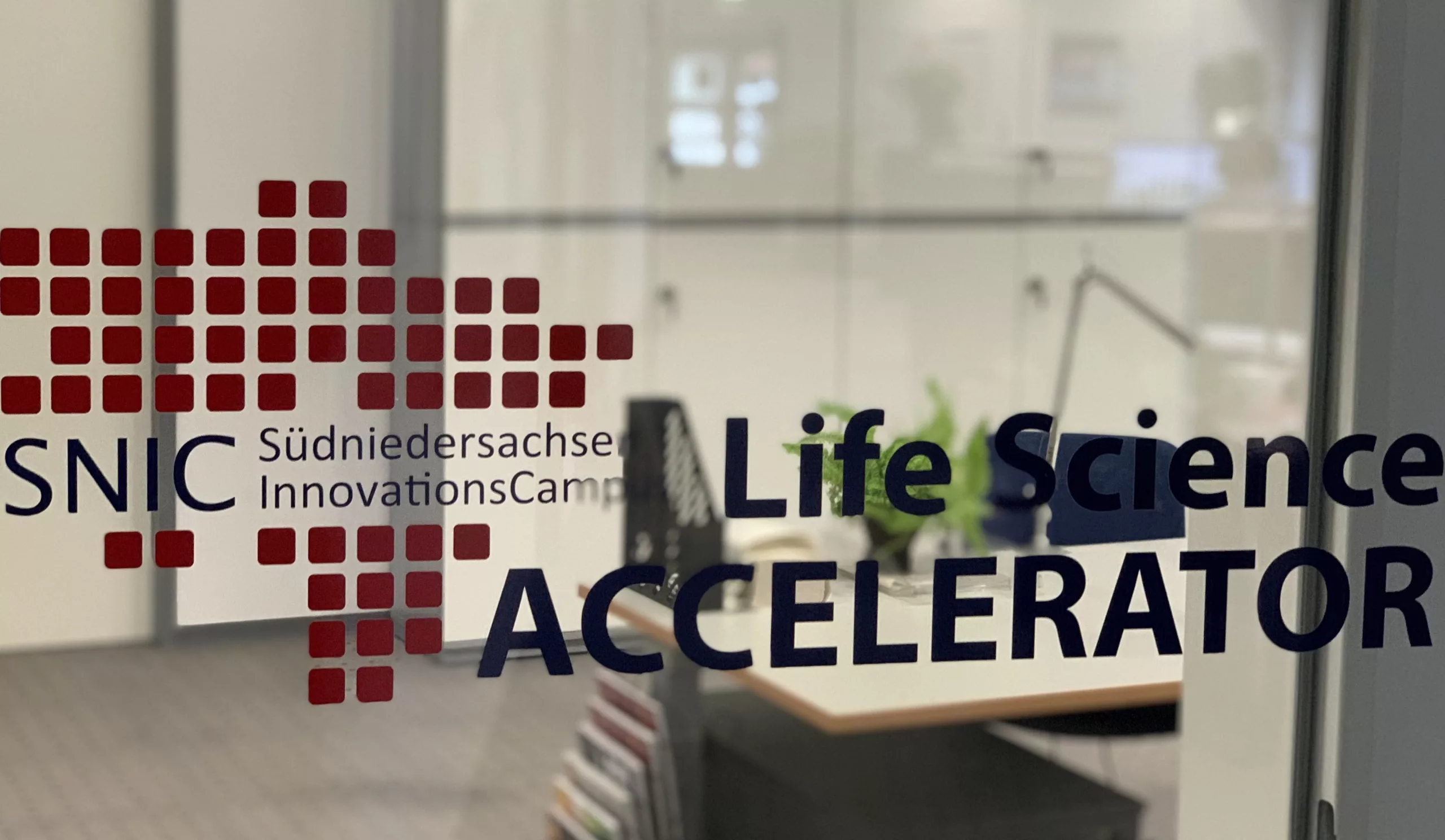Das Bild zeigt eine Glasscheibe mit dem Logo des SNIC Accelerators. Im Hintergrund sieht man ein Büro.