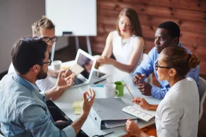 Eine Gruppe von jungen Frauen und Männern sitzt mit Ordner und Laptop am Konferenztisch.