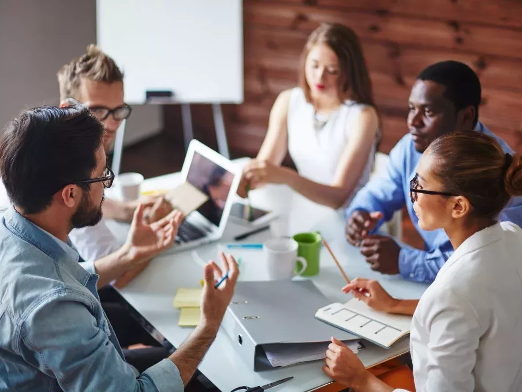 Eine Gruppe von jungen Frauen und Männern sitzt mit Ordner und Laptop am Konferenztisch.