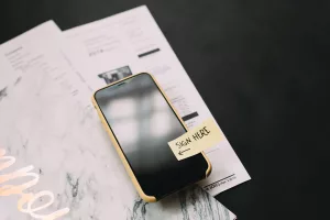 Ein Handy mit einem Post-It "Sign here" liegt auf einem Vertrag.