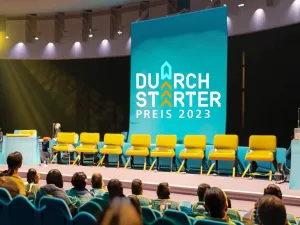 Eine Bühne mit einem Publikum davor. Auf der Bühne steht auf einer Leinwand: Durchstarter Preis 2023
