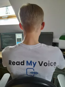 Der Gründer Mika Danner vor seinem Computer von hinten fotografiert. Auf dem Rücken seines T-Shirts ist das Logo seines Startups Read my Voice zu sehen.