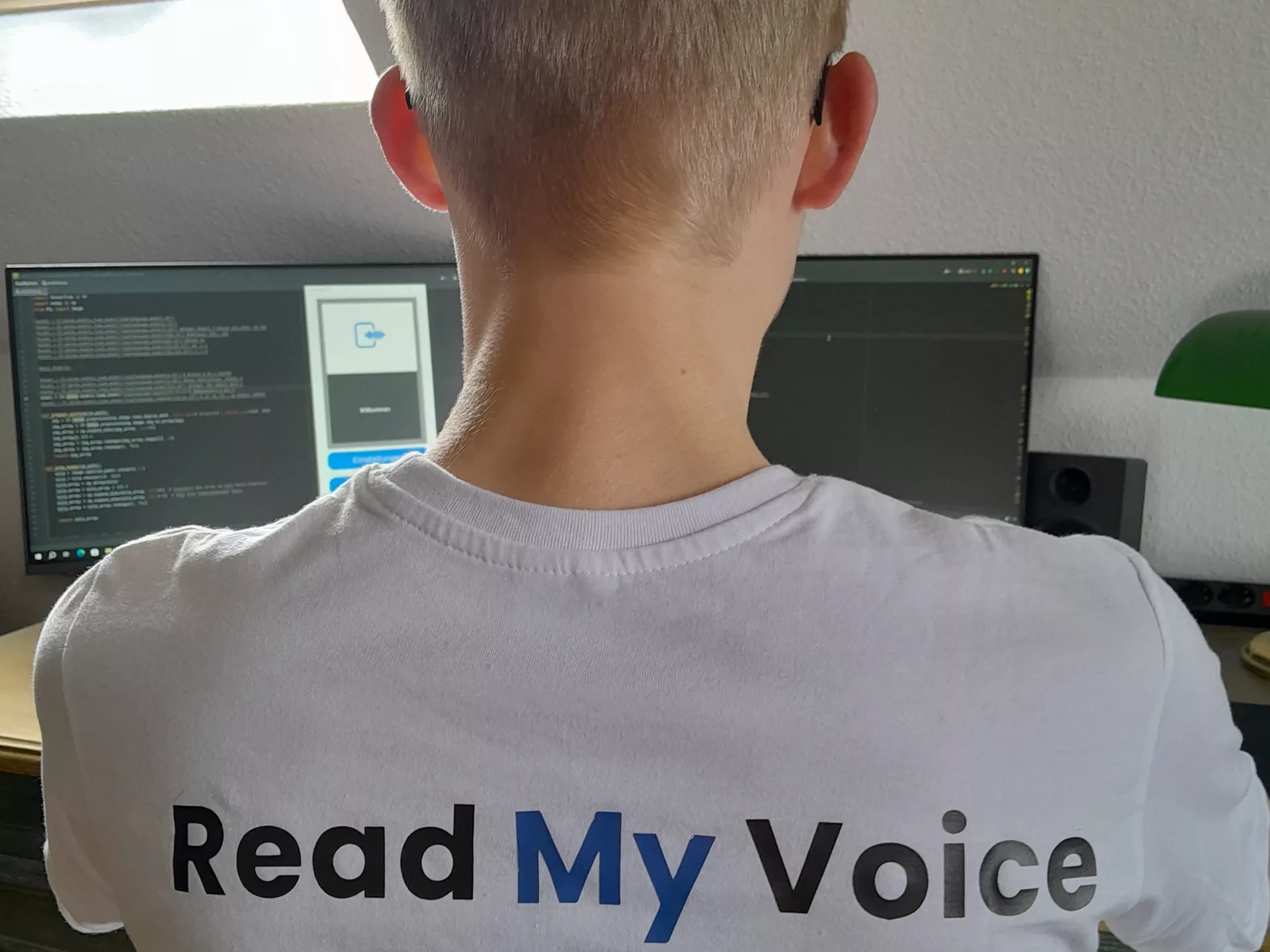 Der Gründer Mika Danner vor seinem Computer von hinten fotografiert. Auf dem Rücken seines T-Shirts ist das Logo seines Startups Read my Voice zu sehen.