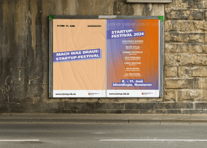 Eine Plakatwand an einer Straße mit zwei Plakaten zum Startup-Festival von Niedersachsen.next Startup. Eins ist orange mit der Aufschrift "Mach was draus: Startup Festival", das andere zeigt das Lineup der Speaker